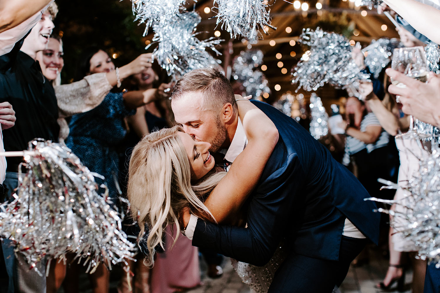 Groom dips bride and kisses her cheek as wedding guests make tunnel around them waving silver pom poms for wedding exit