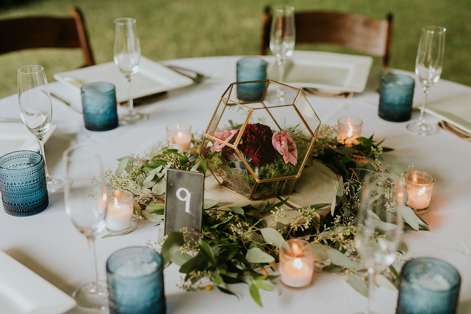 Reception centerpiece with modern gold geometric terrarium vase with moss and individual flowers inside, surrounded by greenery and romantic soft pink votive candles