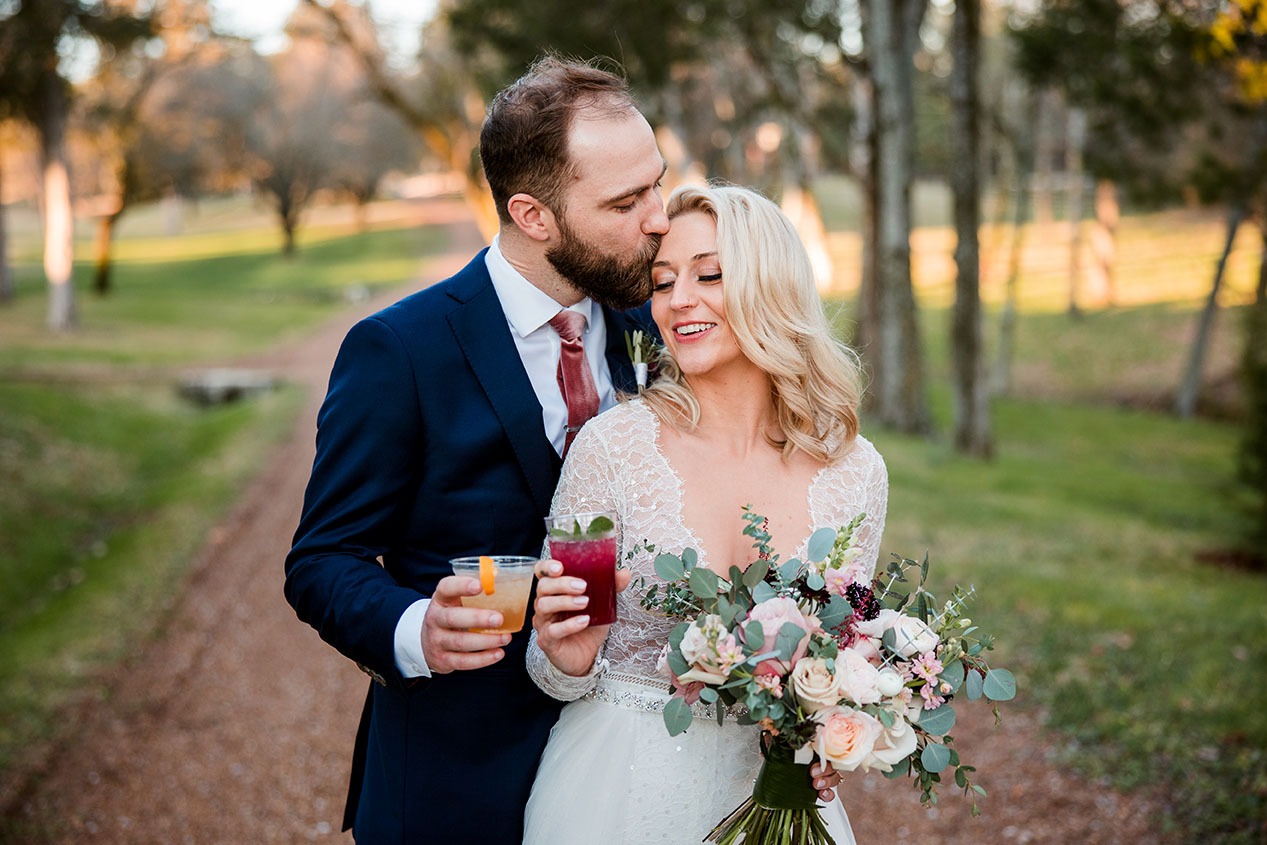 Groom Kissing Top Of Bride's Head While Both Holding Cocktails