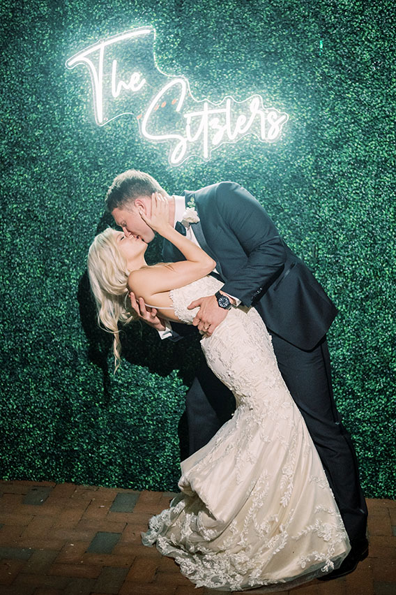 bride and groom kissing in front of neon sign of last name