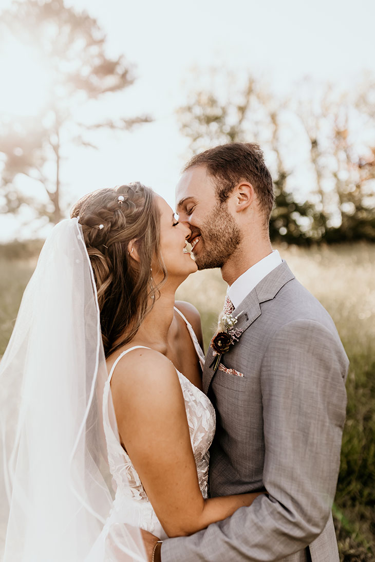 bride with long veil and groom in grey tux kissing at sunset