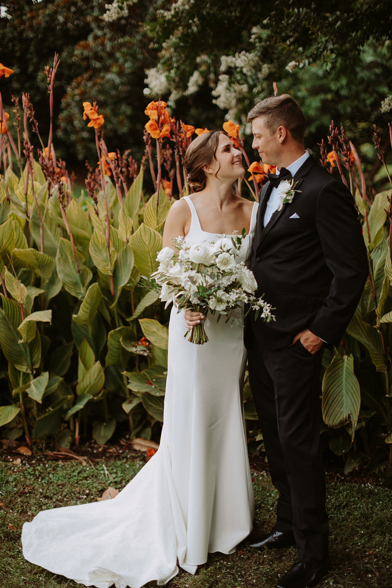 The bride and groom look at each other while smiling during a portrait in front of a garden