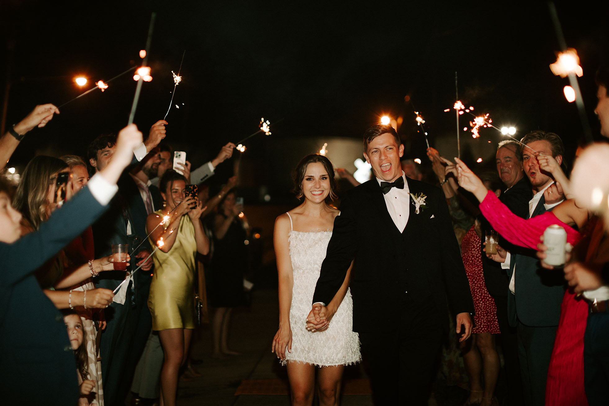 The bride and groom smile as they leave their reception surrounded by guests waving sparklers and cheering