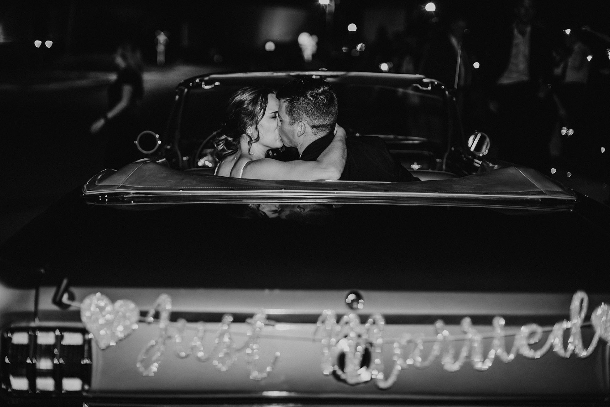 Newlyweds kiss in the backseat of a classic convertible getaway car with a sparkly "just married" sign blurry in the foreground