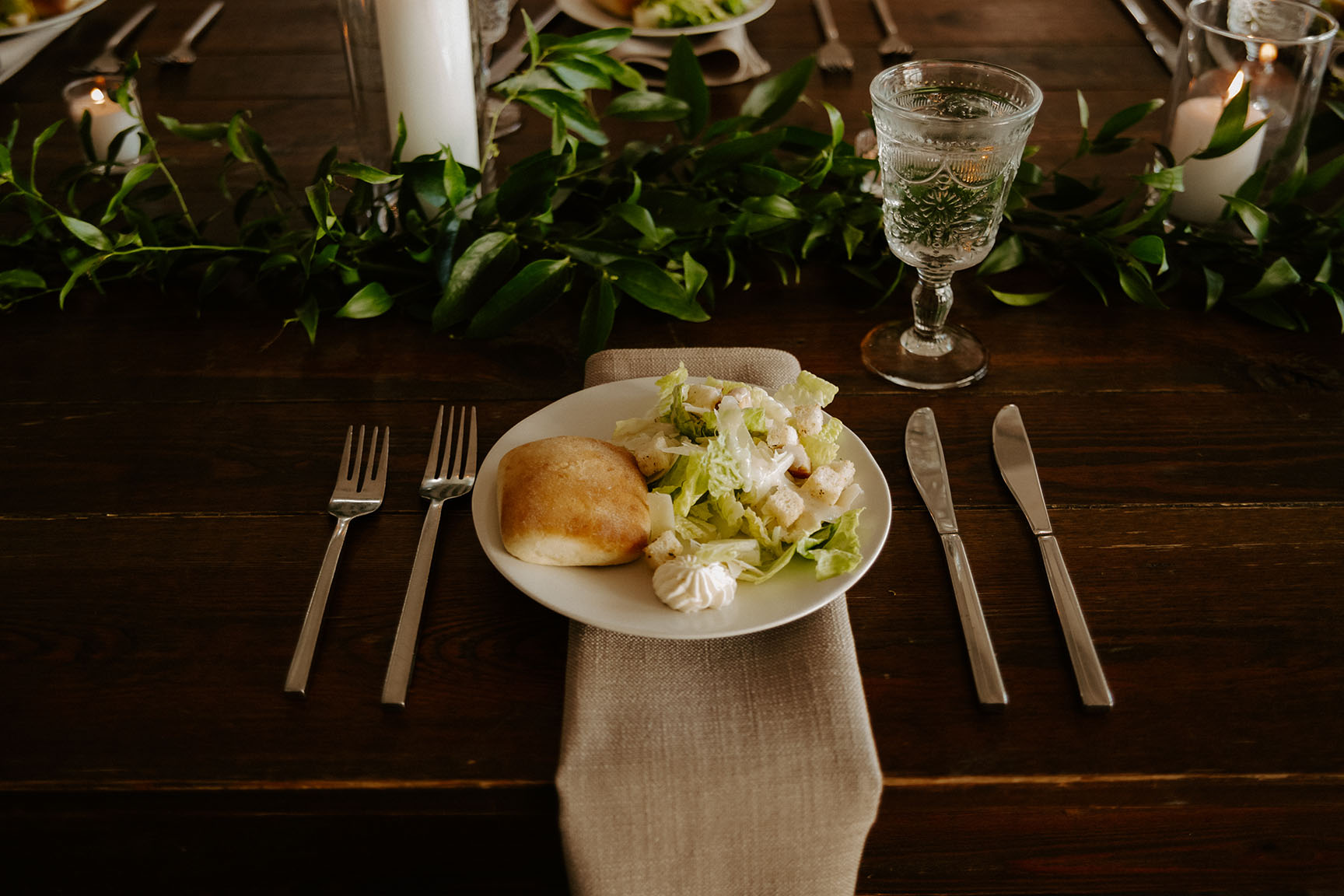 A close-up of a wedding reception place setting with a beige linen napkin, silverware, a pre-set salad, and a greenery garland centerpiece