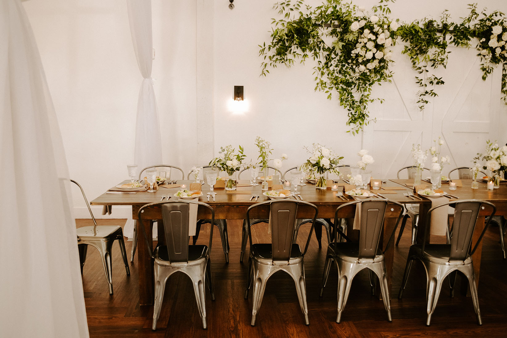 A wooden farmhouse reception table set with metal cafe chairs, beige napkins, and white blooms in small bud vases down the center