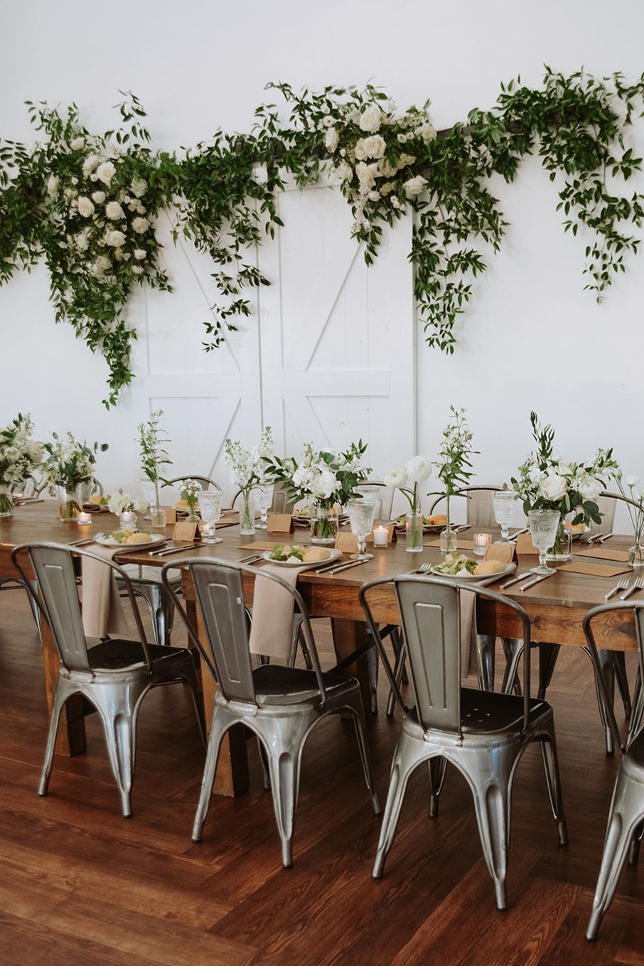 A wooden farmhouse reception table set with metal cafe chairs, beige napkins, and white blooms in small bud vases down the center