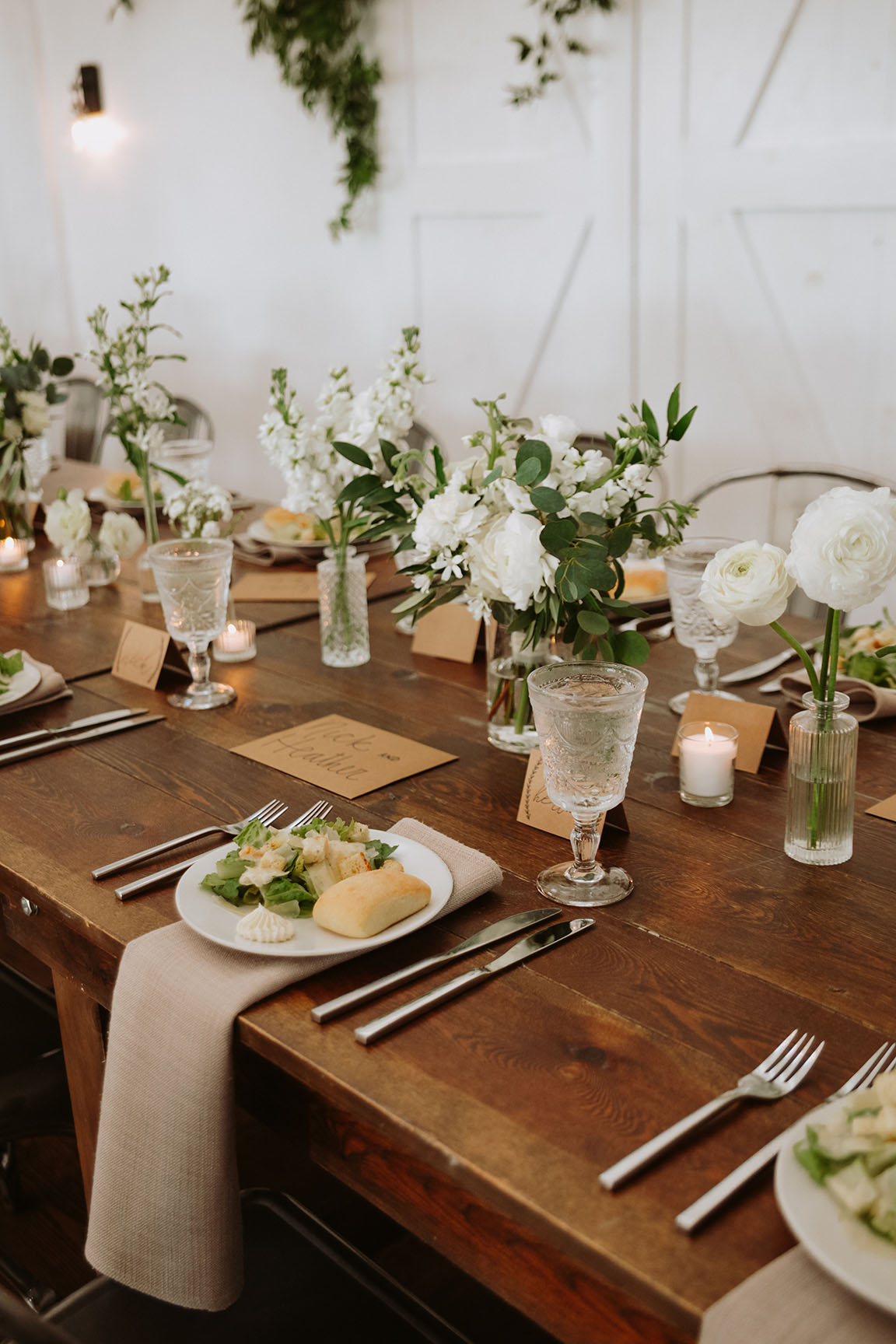 A close-up of a wedding reception table setting with a pre-set salad, beige napkin, brown paper name card, and white blooms in small bud vases down the center