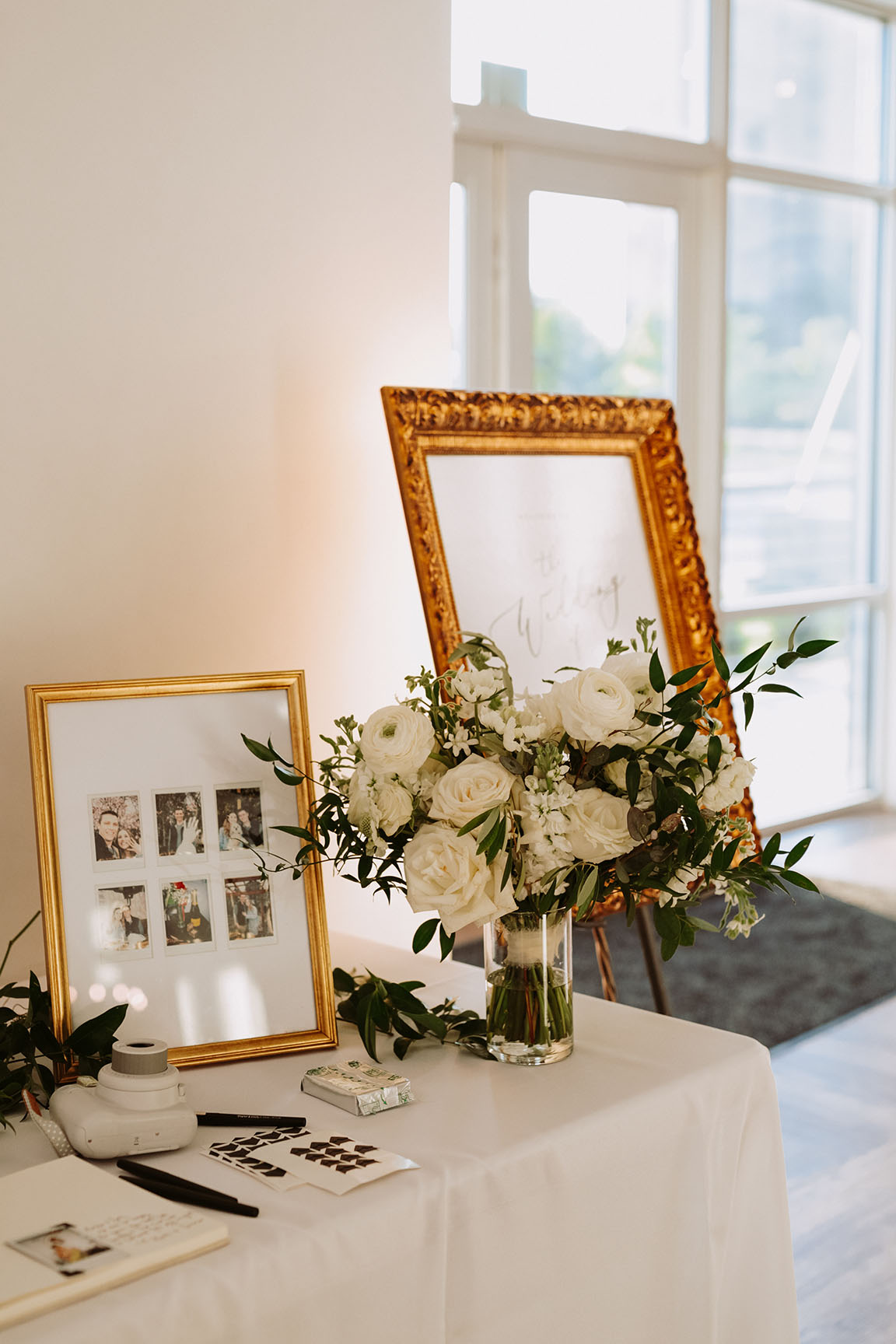 The corner of a wedding reception table with a white floral arrangement in a glass vase, a framed collage of photos of the bride and groom, and an instant camera for their photo guest book