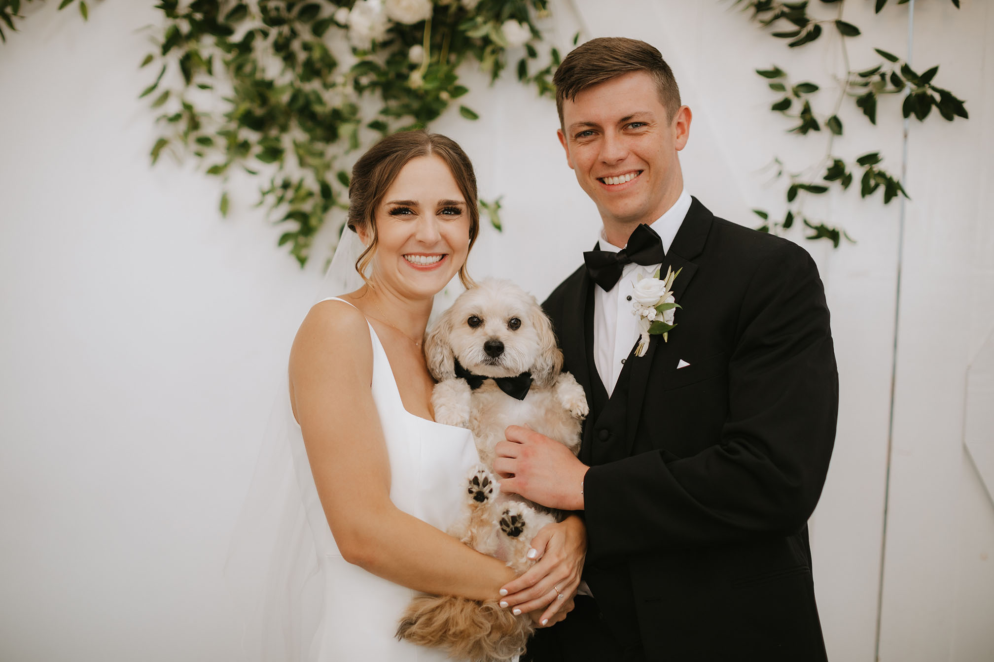 The bride and groom pose with their small white dog that is dressed up with a black bowtie for the wedding