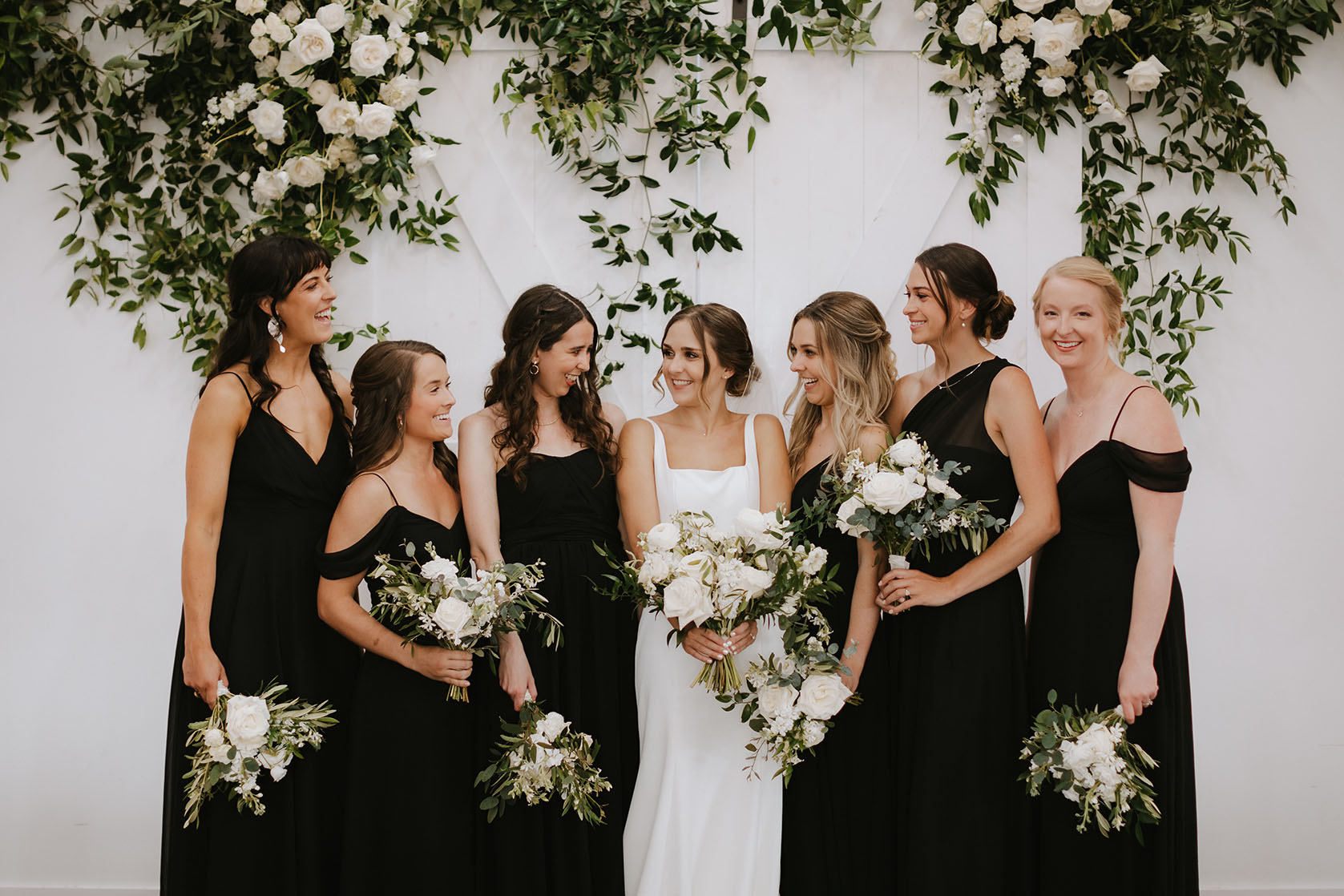 The bride poses with her wedding party, all dress in different styles of black dresses with white and green bouquets