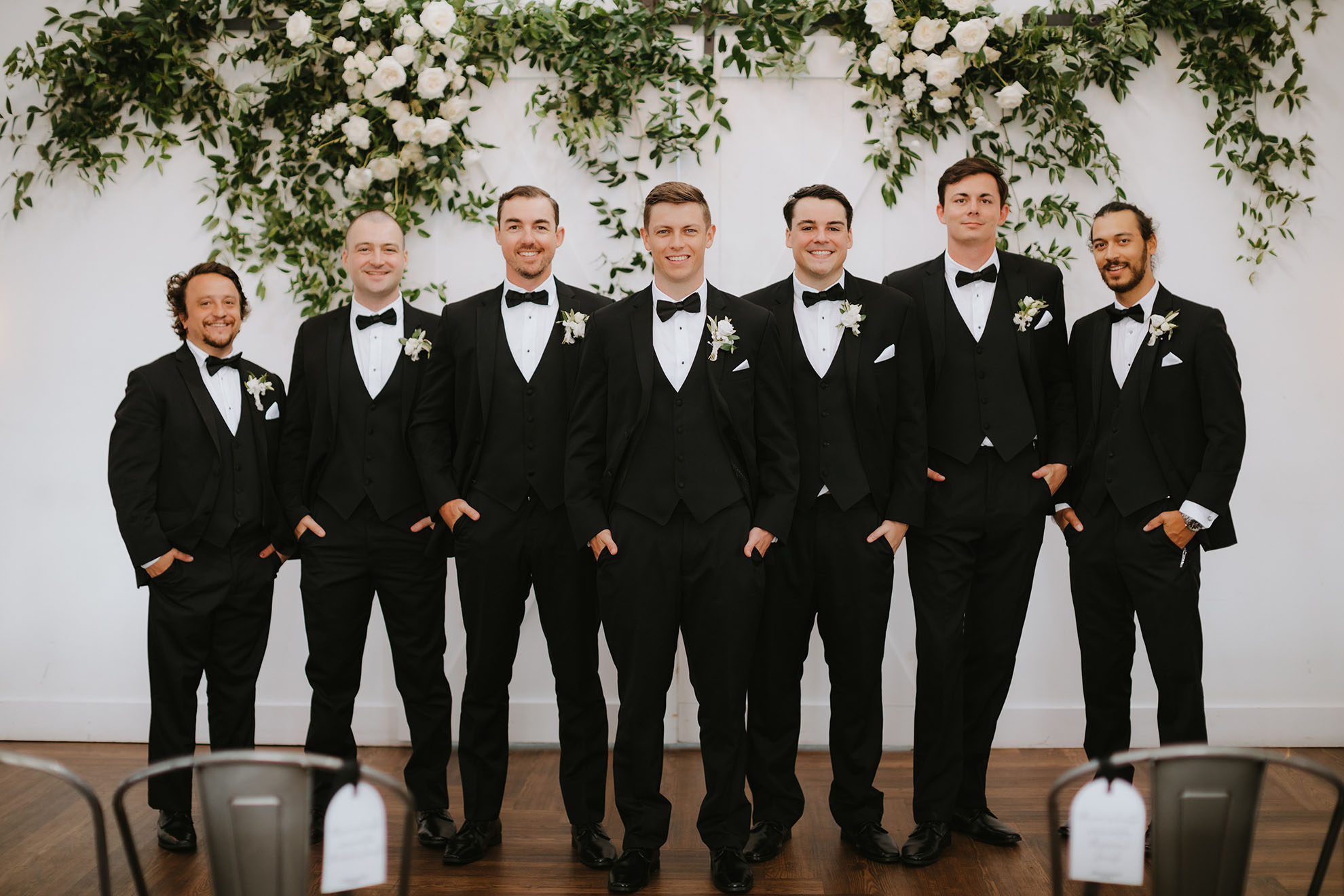 The groom poses with his wedding party, all in classic black three-piece tuxedos with their hands in their pockets