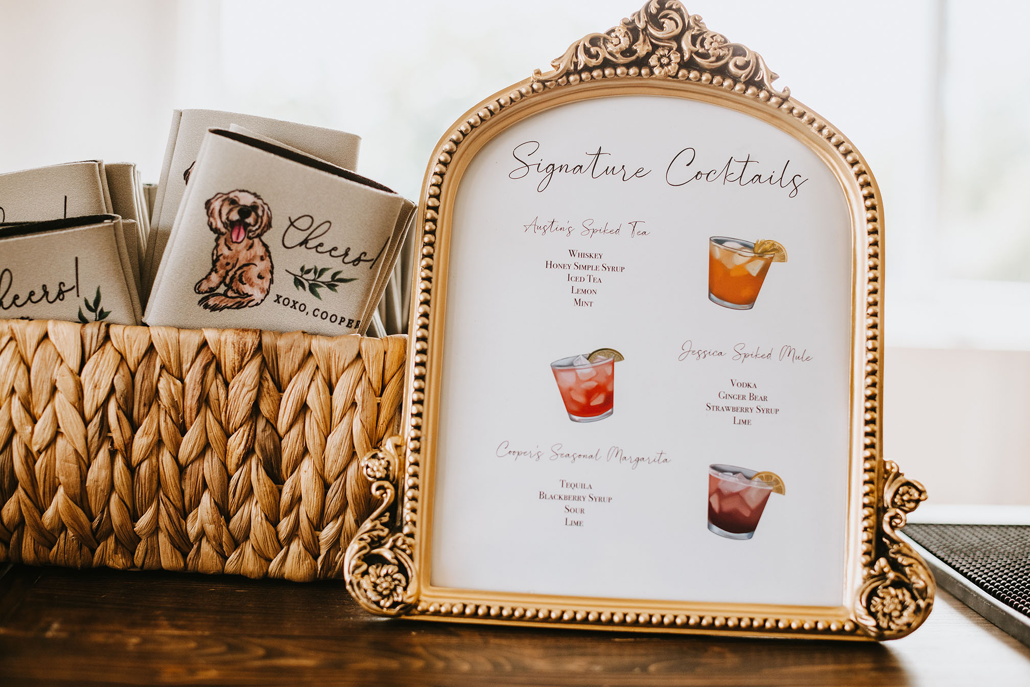 A custom illustrated signature cocktail bar sign sitting next to a basket full of customized drink koozies