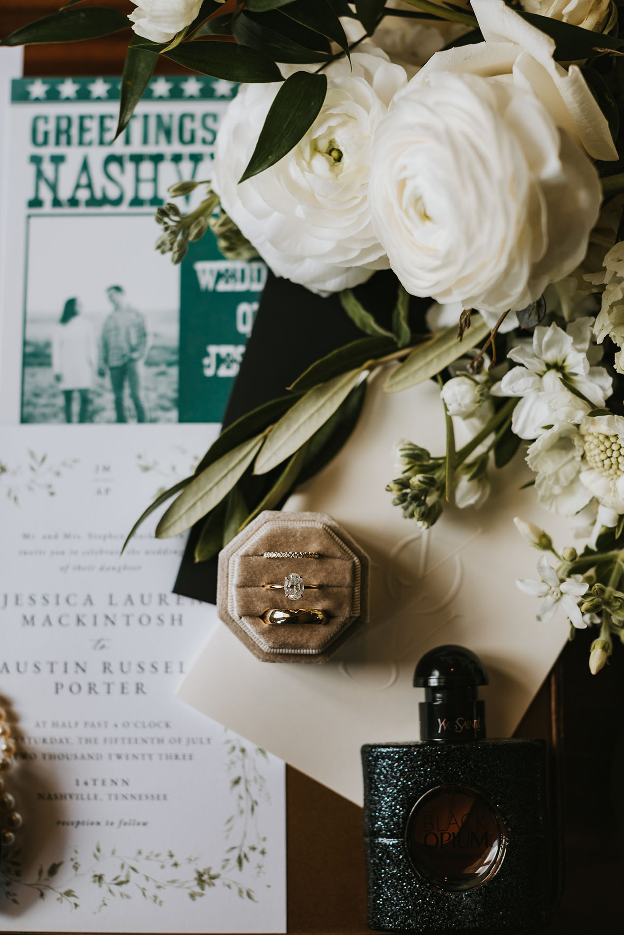 A flat lay photo of wedding day details, including their invitation suite, a champagne velvet ring box with their wedding bands, the bride's bouquet, and a bottle of Yves Saint Laurent perfume