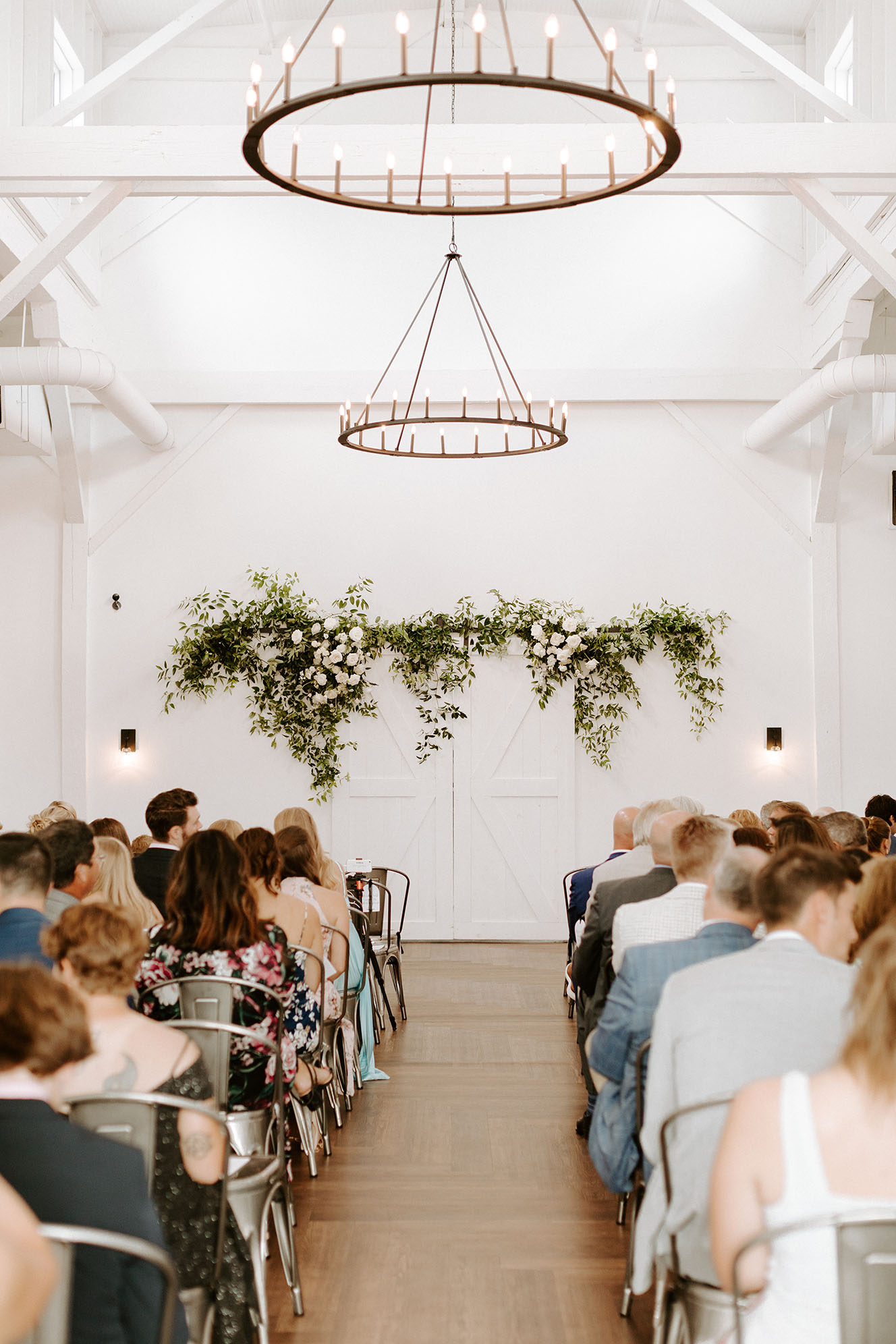 A view from the back of the ceremony aisle toward the altar area with a greenery installment hanging above, as guests look ahead waiting for the ceremony to begin