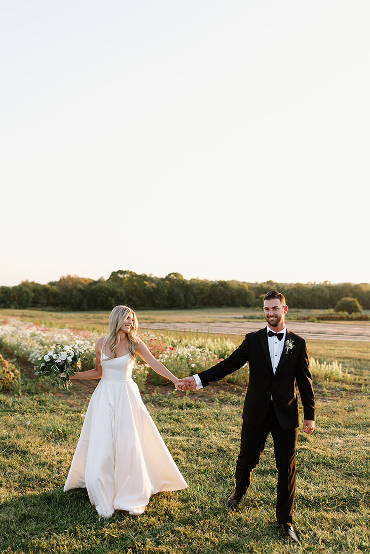 Bride and groom hold hands and smile while walking through farm field at sunset