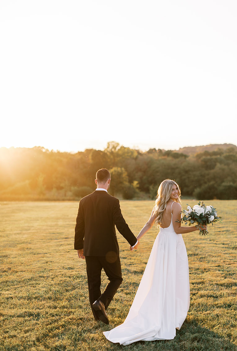 Bride holds hand with groom while walking through farm field at sunset