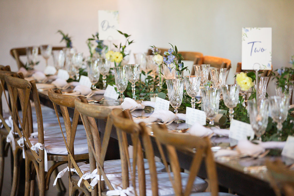 Dark wood farmhouse table decorated with greenery runners, textured crystal glassware, knotted white napkins, and crossback chairs