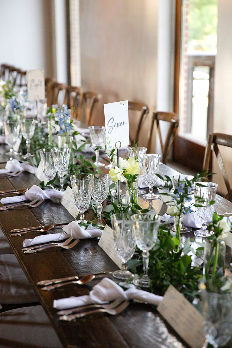 Dark wood farmhouse table decorated with greenery runners, textured crystal glassware, knotted white napkins, and crossback chairs