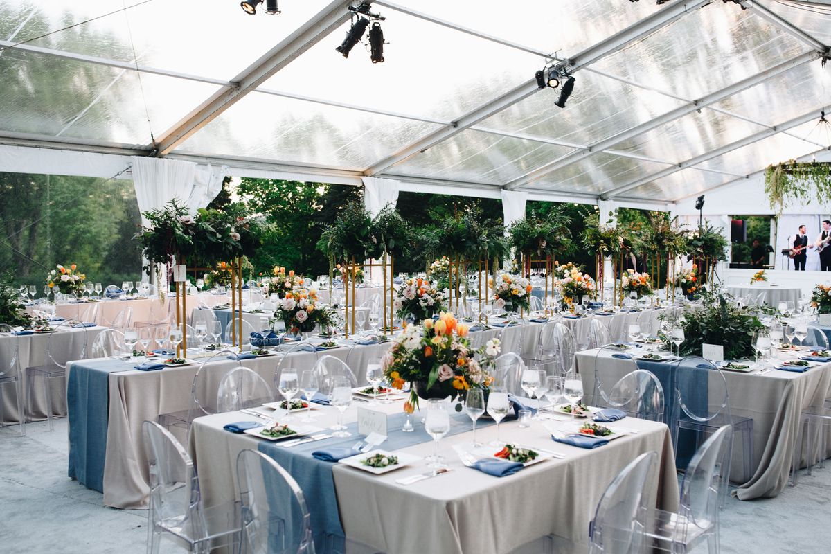 A wide shot of a modern and Southern wedding reception table setup with blue and white linens, a plated Caprese salad, clear acrylic chairs, and centerpieces of peach and pink flowers with greenery