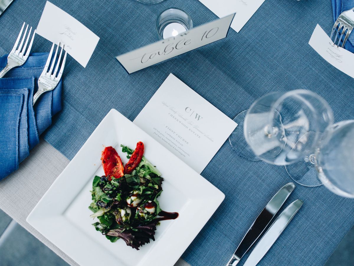 A close-up bird's eye view of a wedding reception place setting with blue linens, a plated Caprese salad, and a custom menu card