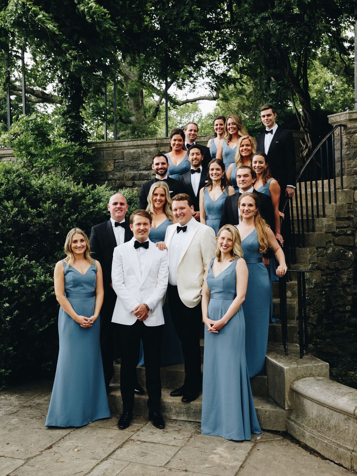 Two grooms pose with wedding party with groomsmen in classic black tuxedos and groomsmaids in dusty blue dresses