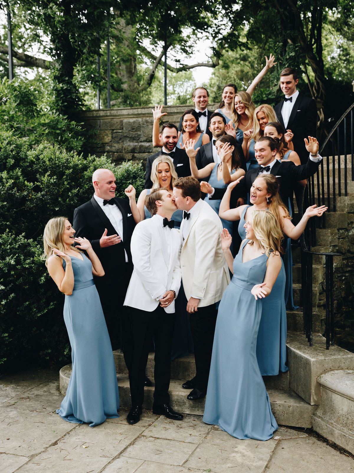 Two grooms pose with wedding party with groomsmen in classic black tuxedos and groomsmaids in dusty blue dresses