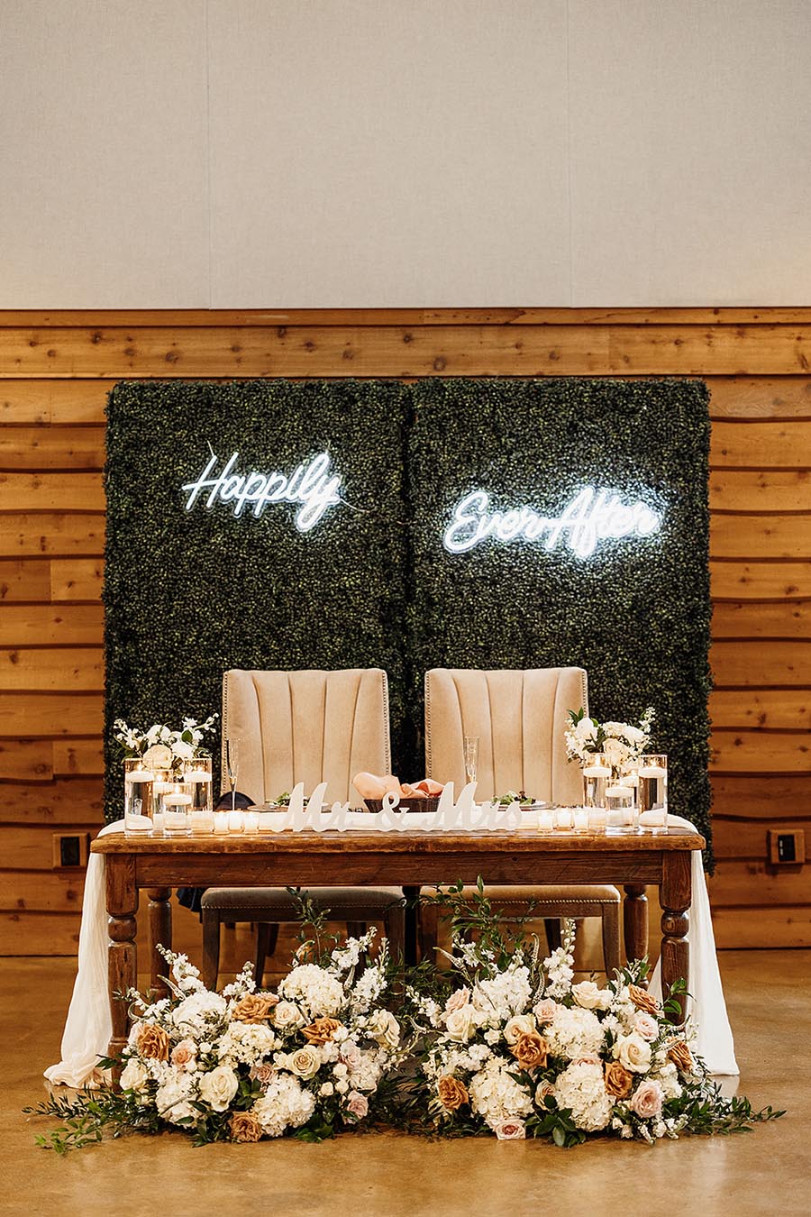 Sweetheart table setup with greenery wall backdrop, custon neon sign that reads "happily ever after," beige velvet chairs, a wooden table with a gauze runner, and large arrangements of neutral flowers with whimsical greenery