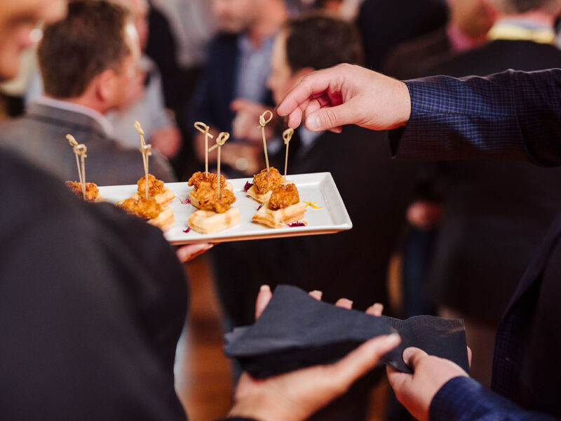 A close-up of a guest reaching for a chicken and waffle bite on a white plate being held by a cater water