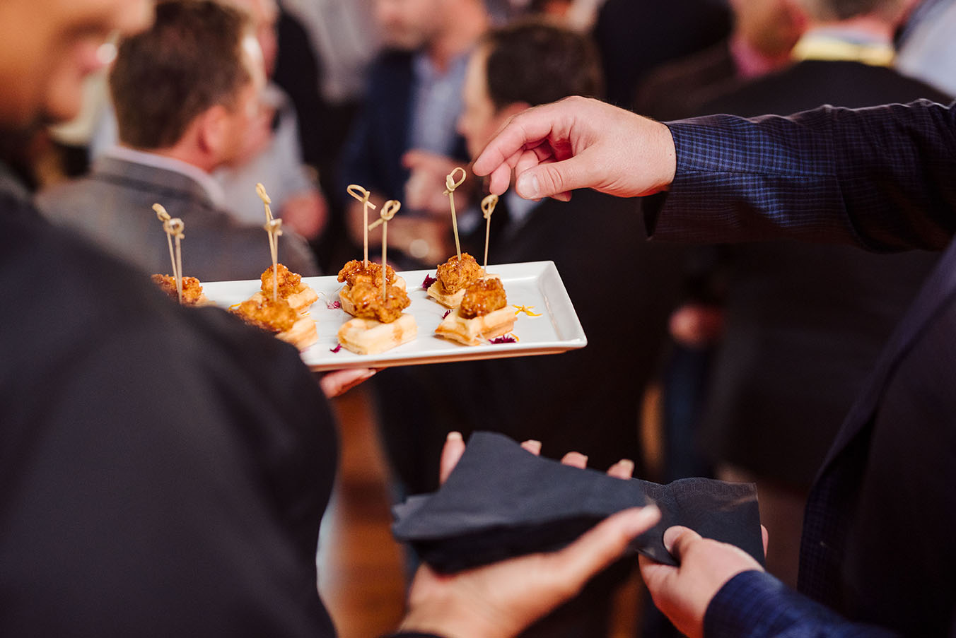 A close-up of a guest reaching for a chicken and waffle bite on a white plate being held by a cater water