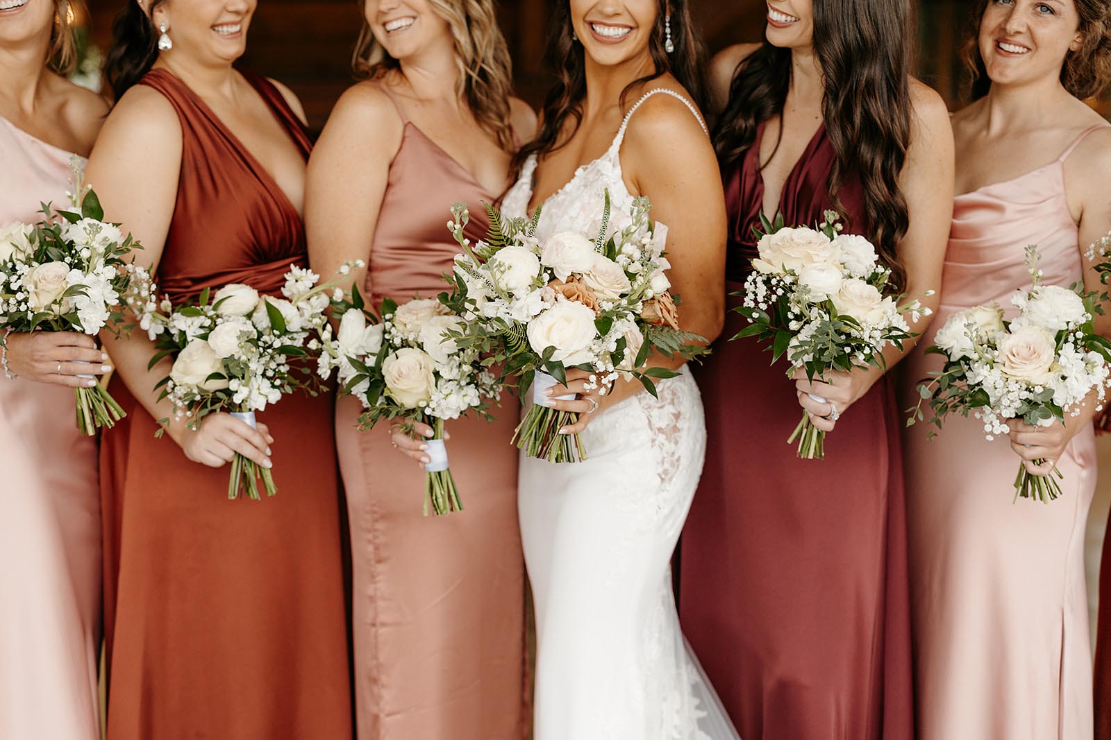 A cropped photo of a bride with her bridesmaids in various shades of red and pink dresses holding all-white bouquets with greenery