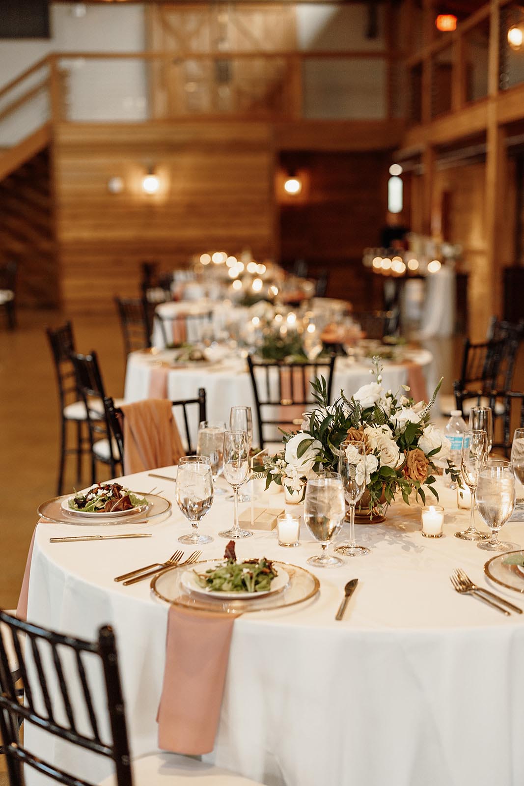 A wedding table set up in a barn with white linens, blush pink napkins, a preset salad on a glass gold-rimmed charger, and a white floral centerpiece with greenery.