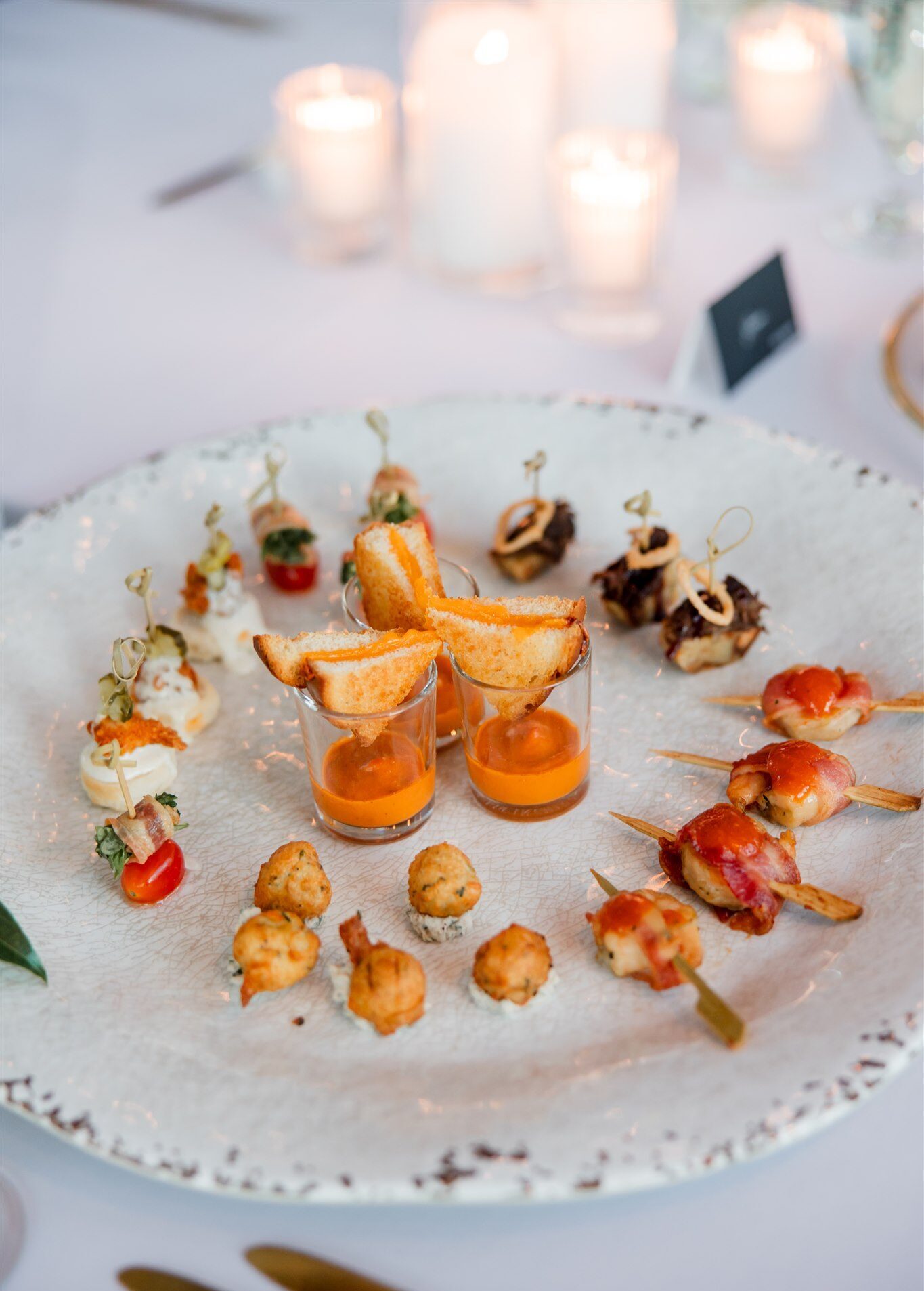 Plate of wedding appetizers including mini grilled cheese with tomato soup shooters, bacon-wrapped skewers and fritters on a candlelit table