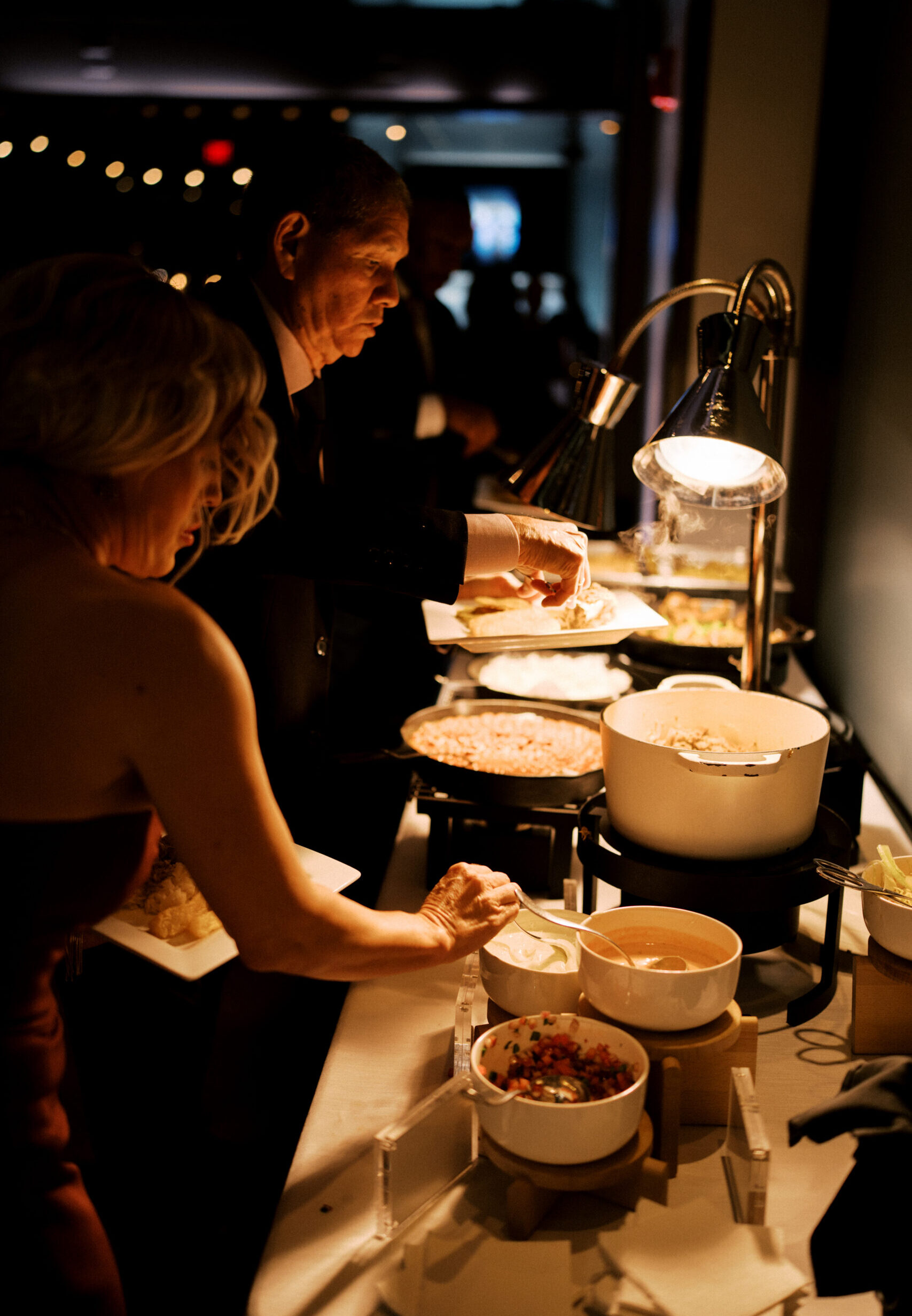 Wedding guests serving themselves from a warm buffet under heat lamps at an evening reception