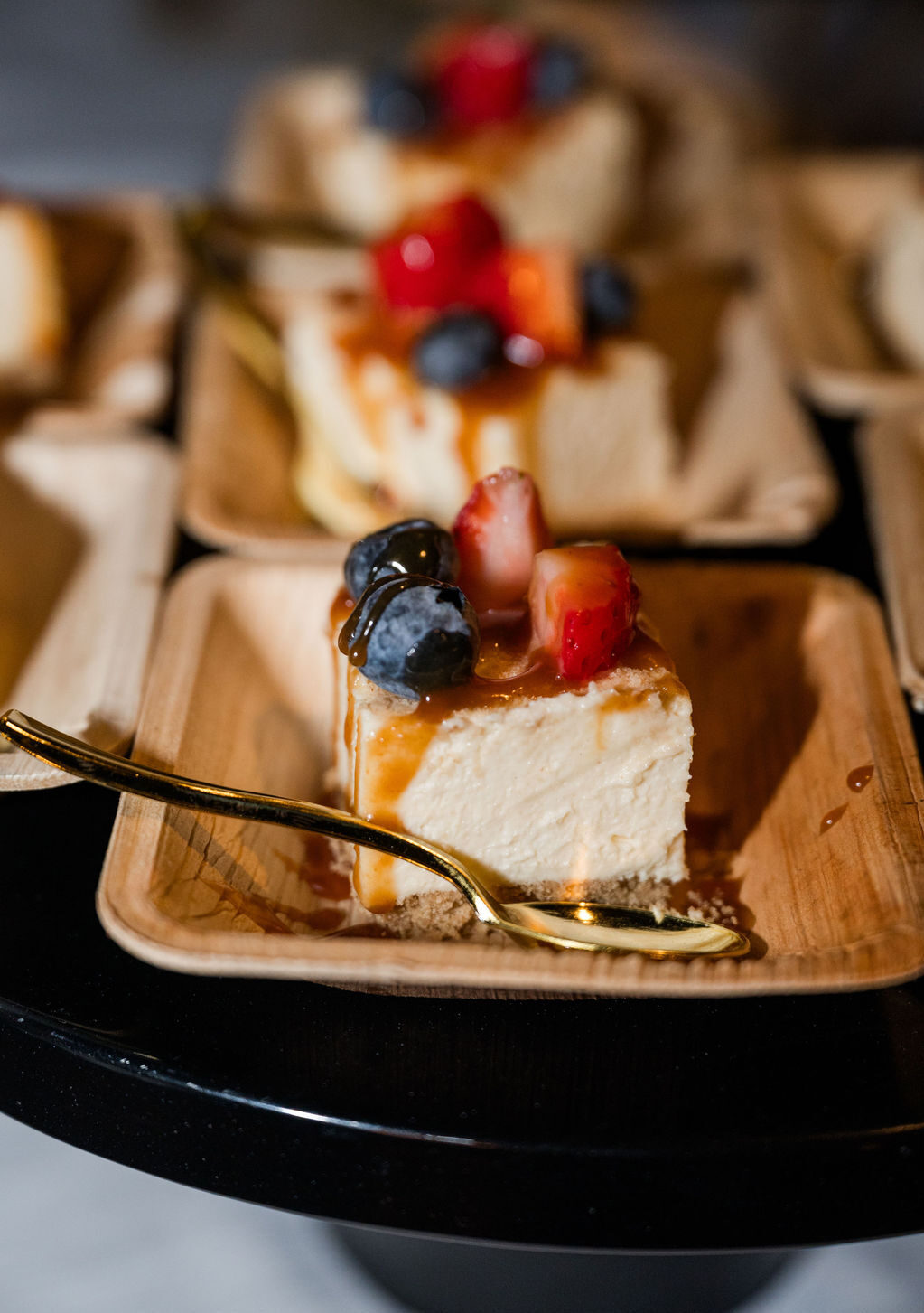 Slice of cheesecake topped with strawberries, blueberries and caramel drizzle on a palm-leaf plate with a gold spoon