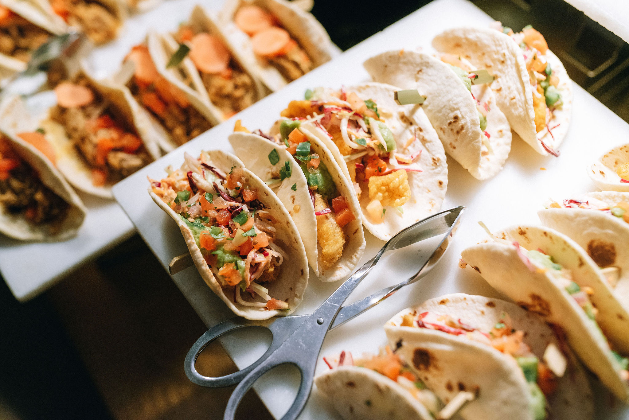 Mini fish tacos with slaw, guacamole and fresh pico de gallo lined up in taco holders with serving tongs
