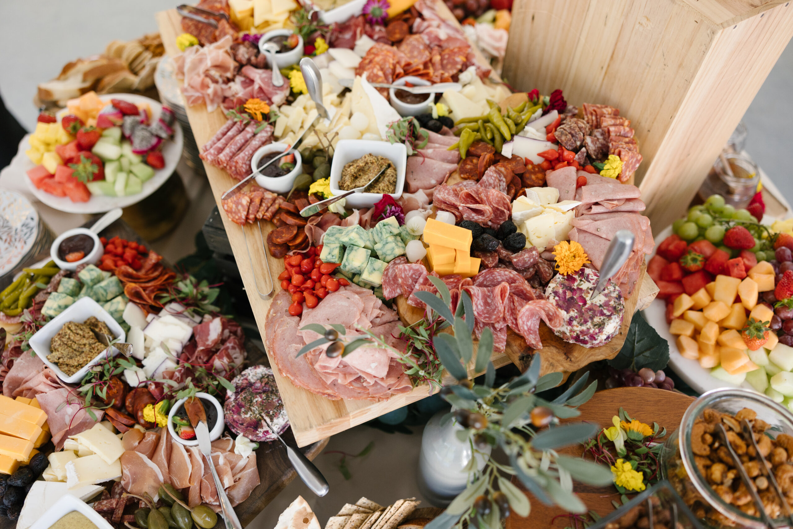 Grazing table with cured meats, assorted cheeses, whole-grain mustard, peppers and edible flowers on wooden boards, with fresh fruit platters alongside