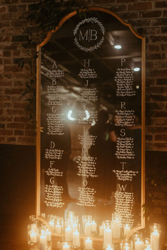 Seating chart in white calligraphy on tall mirror with ornate gold frame, surrounded by pillar candles in hurricane vases